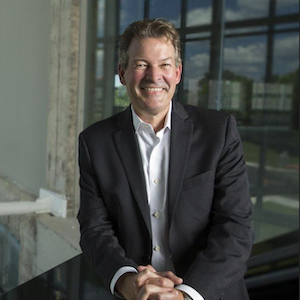 David Adams standing against a railing in front of a large glass window, wearing a dark suit jacket with a white shirt