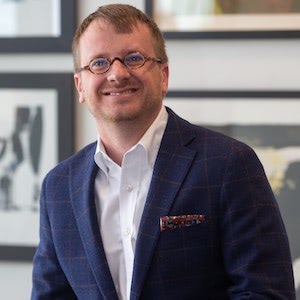 Ryan Hays, standing in front of a wall covered in framed pictures, wearing a dark blue suit with a white shirt