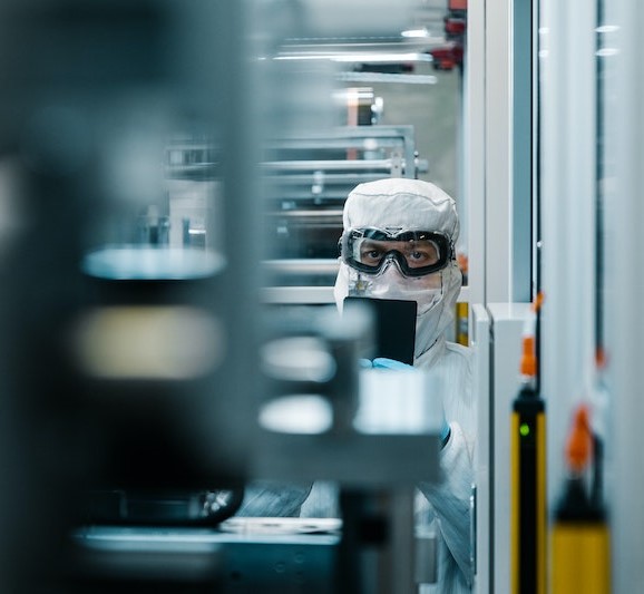 Man in white protective gear and black goggles standing with handheld device in factory