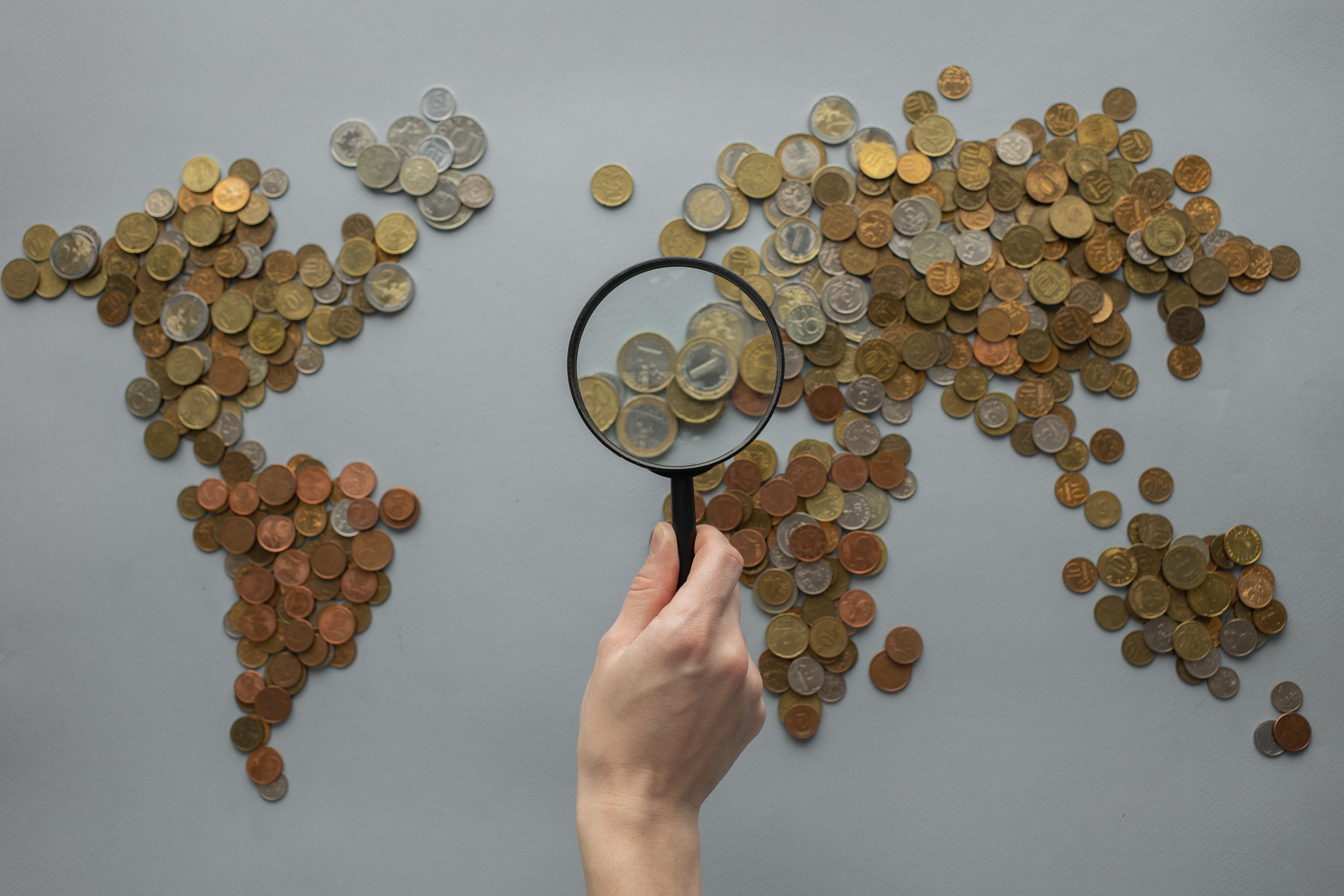 a world map laid out in coins, with hand holding a magnifying glass over the coins representing continental Europe