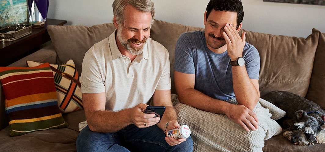Two men sitting on couch while one looks at medication instructions on his phone