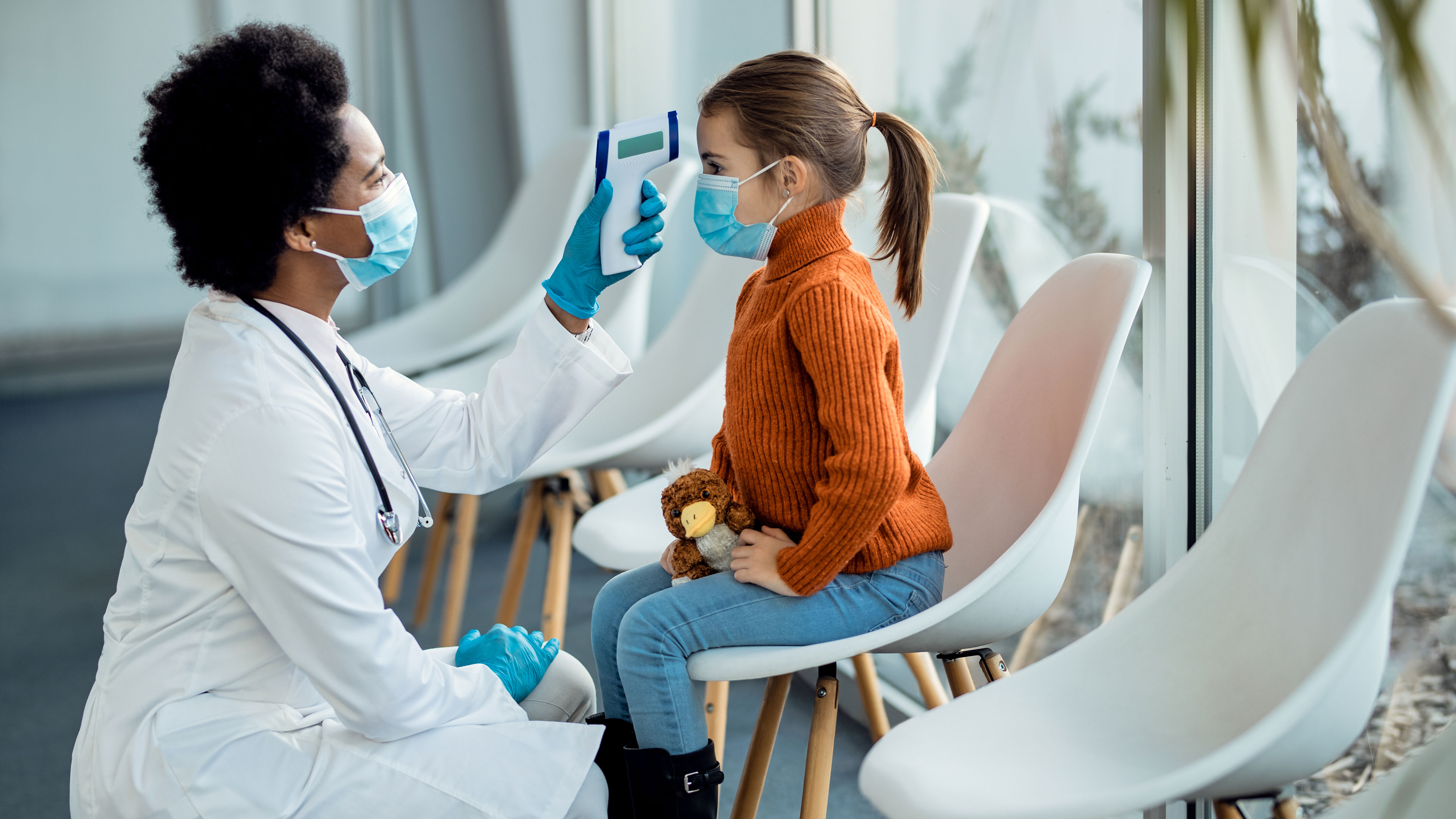 Masked female doctor takes temperature reading of masked girl in waiting room