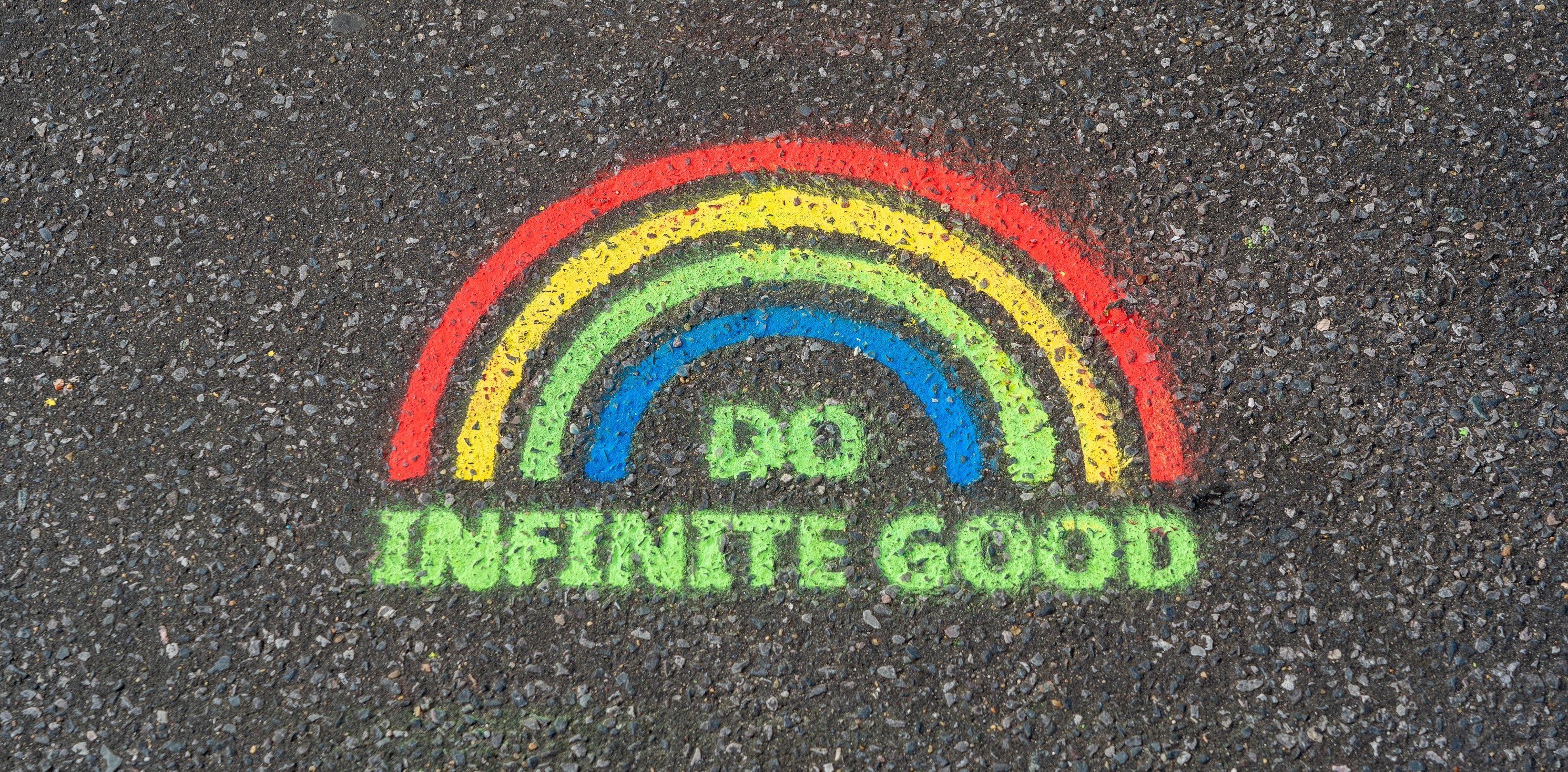 a stencil graffiti of a rainbow with the words “do infinite good" in all caps written underneath, spraypainted onto tarmac