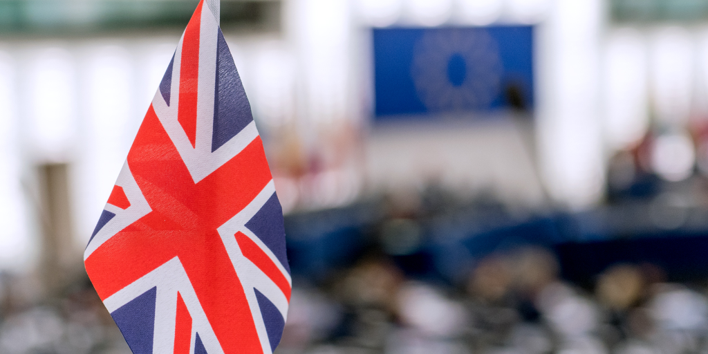 a small UK flag standing on a desk in the EU Parliament, with the EU flag blurred in the background of the plenary