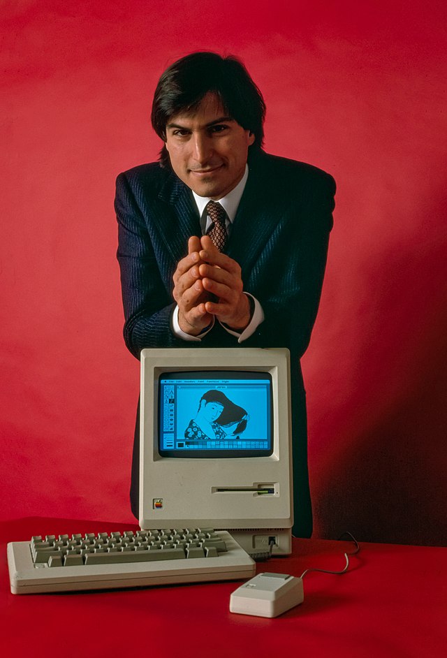 Steve Jobs leaning on Apple Macintosh in front of red background, circa 1984