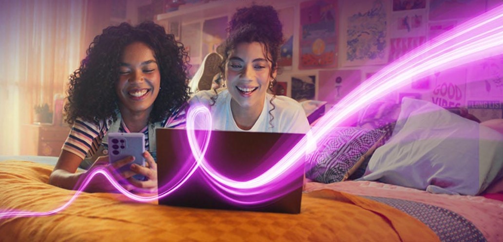 Two teenage girls on a bed looking at a phone and a laptop with a pink light trail in front of them