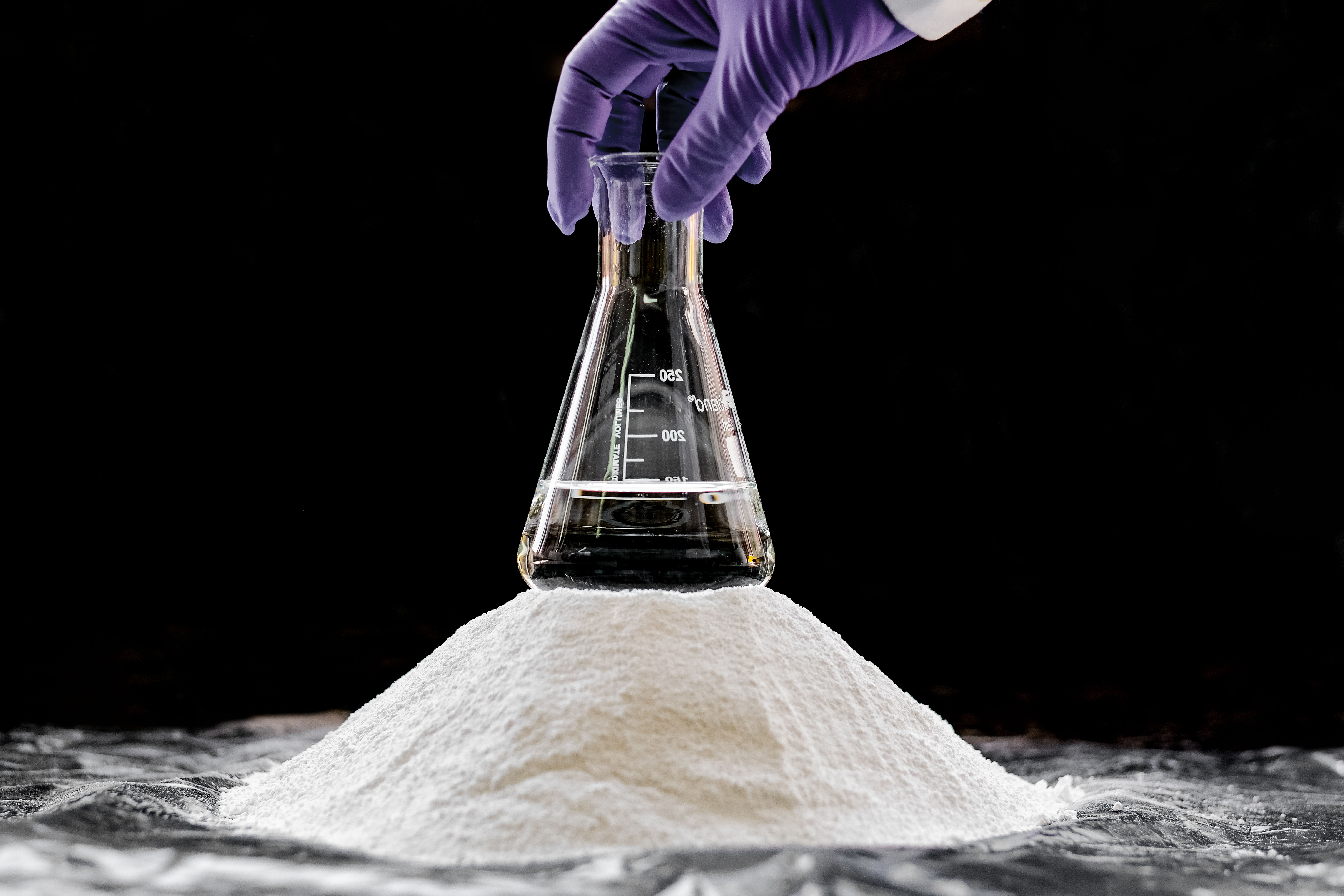 Purple gloved hand holds conical flask of liquid on top of mound of white powder against black background