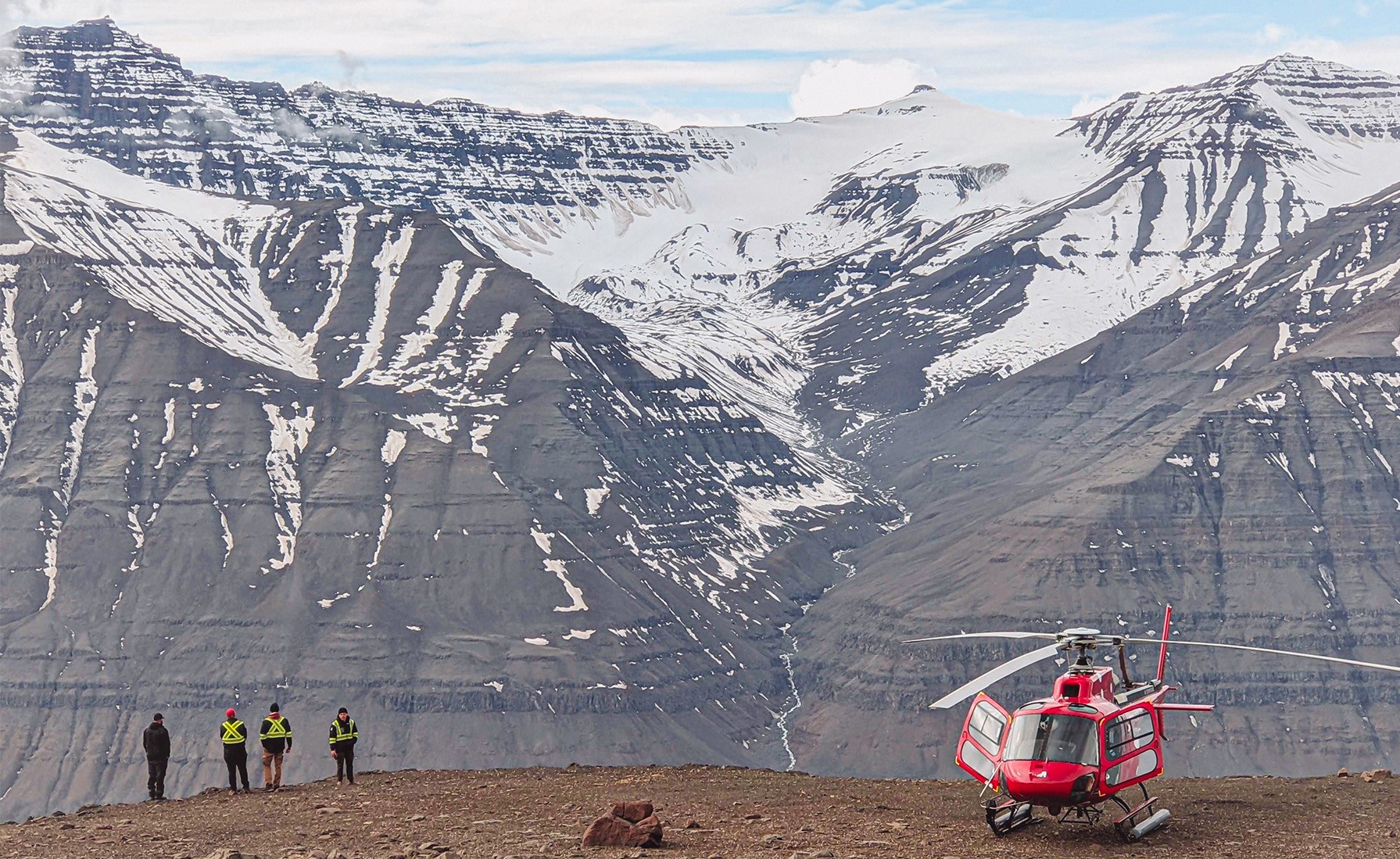 Four people standing next to a red helicopter on a ridge looking at a snow-capped mountain