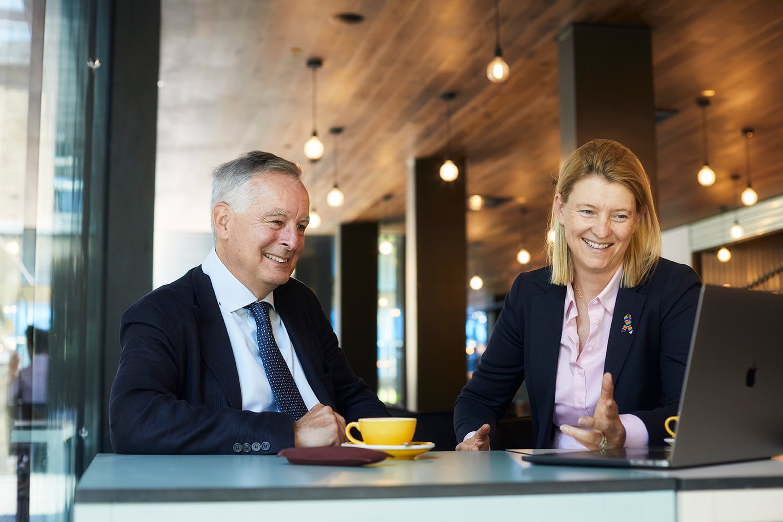 Mike Rees (right) and Lisa Smith (left) sitting in front of and looking at a laptop. There is a yellow cup in front of Mike Rees. Both of them are smiling.