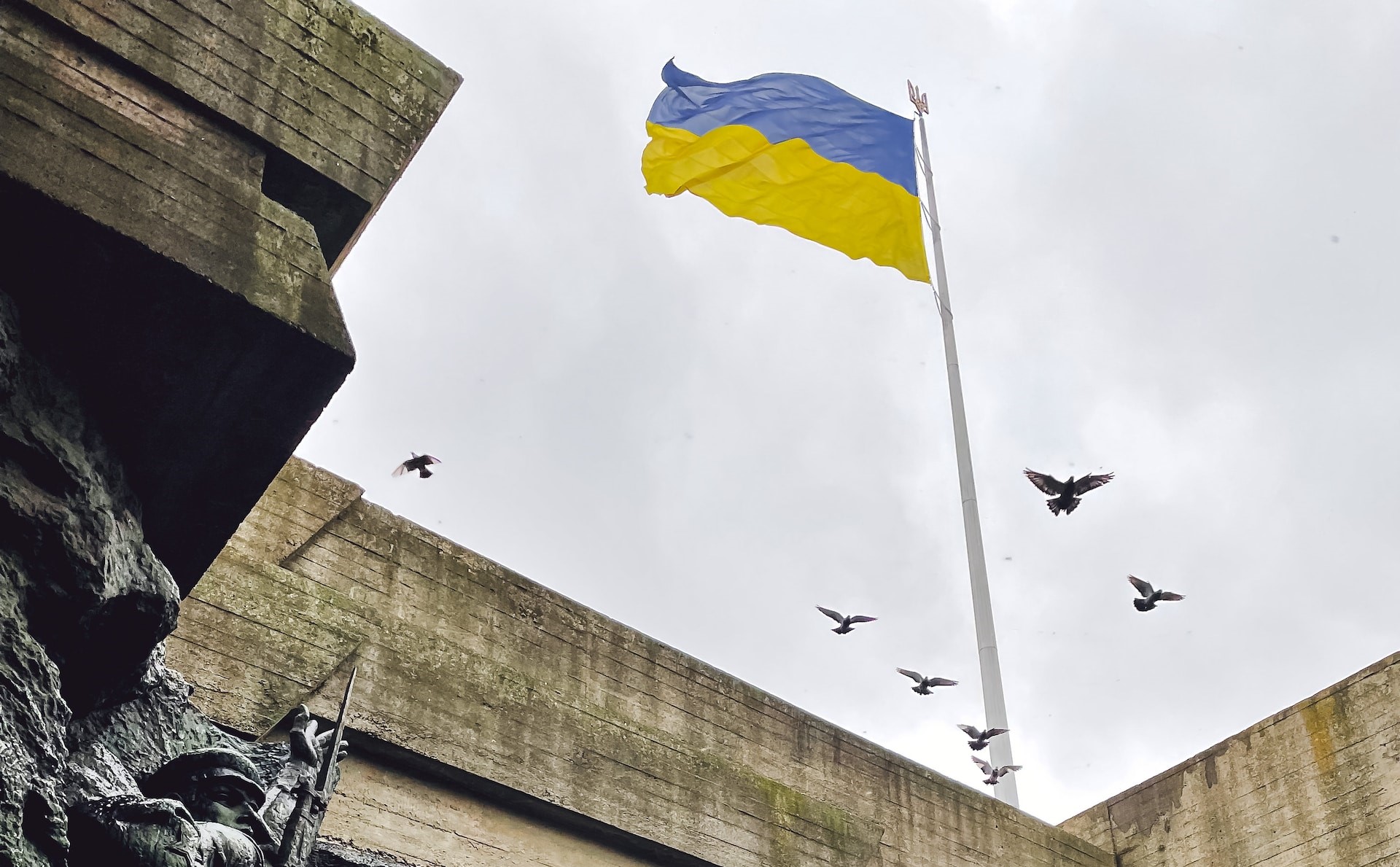 A Ukrainian flag flying over a war memorial as birds fly up