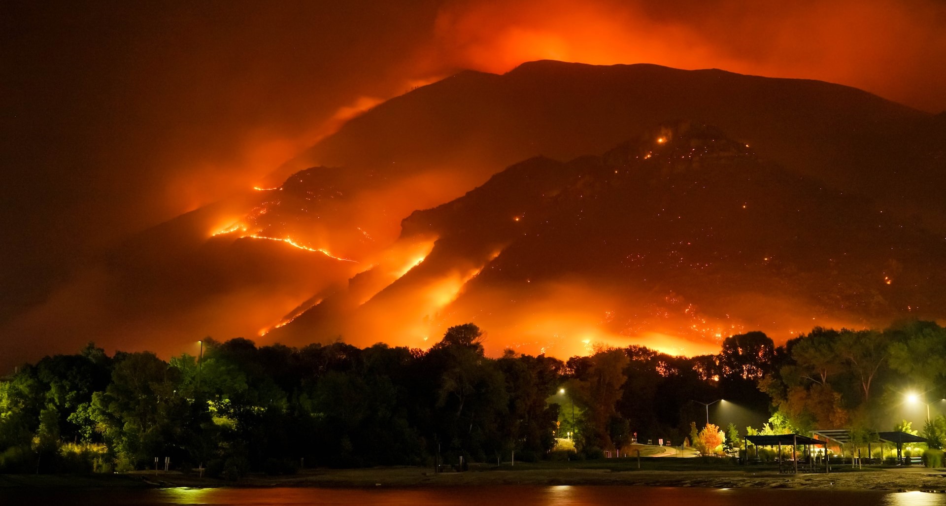 Forest fire engulfs a mountain at night overlooking a forest and river settlement below