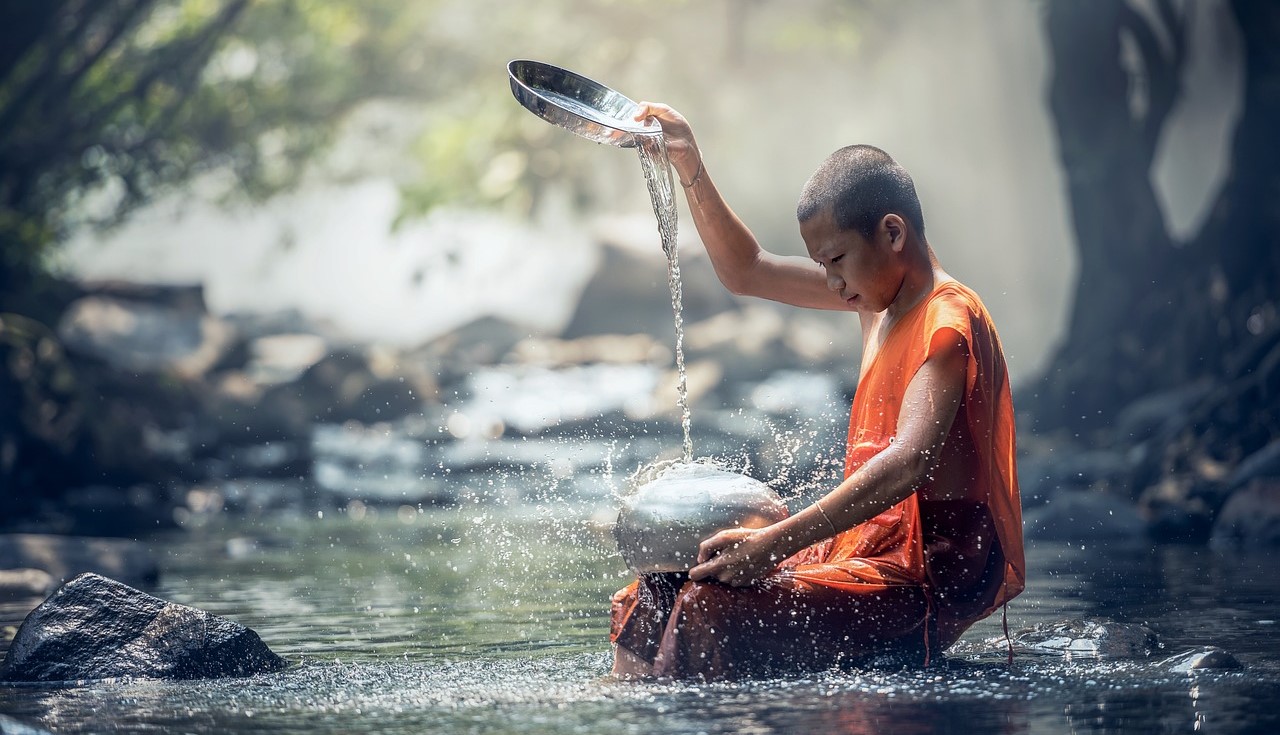 Boy in orange monk's robes sits in river, pouring water from a dish onto a silver object