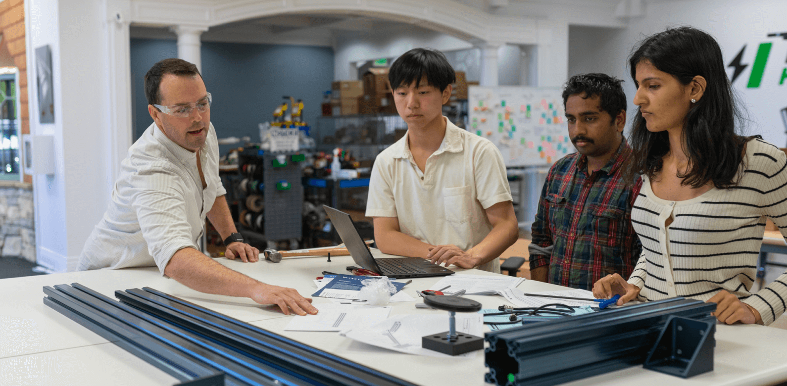 A man in lab goggles point to a piece of paper on a table while three younger workers look on