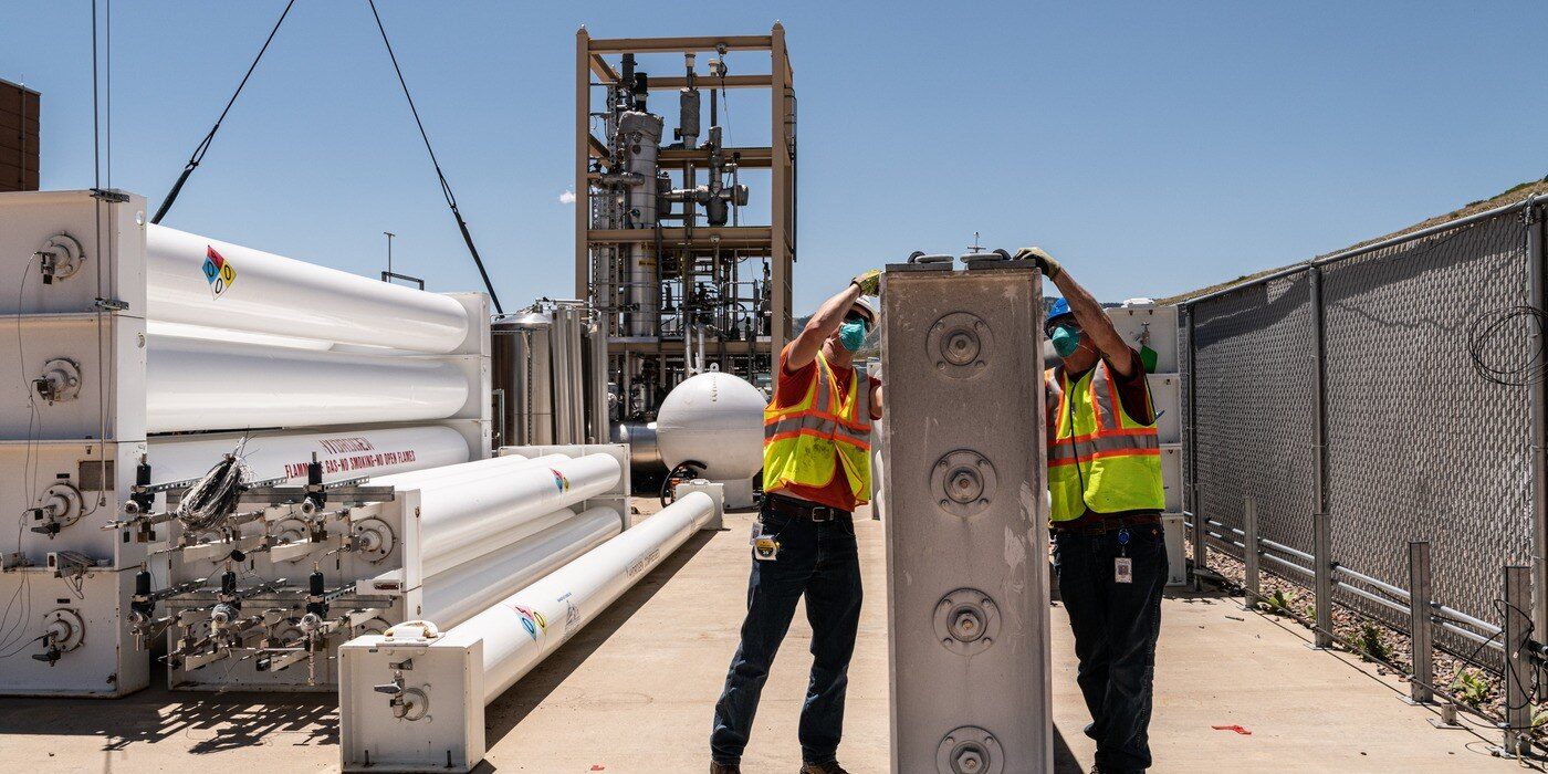 Two construction workers handle a large concrete component in front of a hydrogen electrolyser