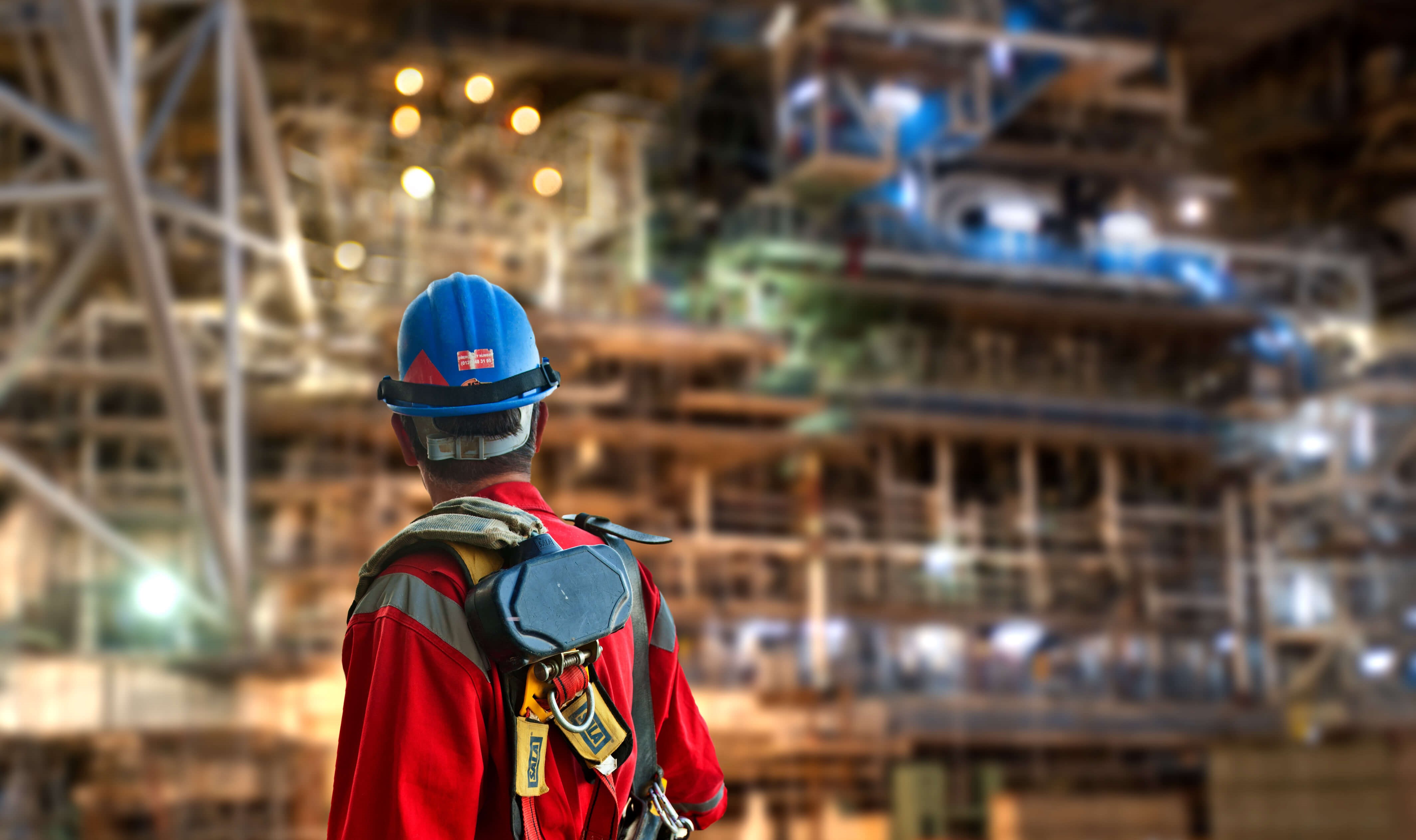 Man in red boiler suit and blue hard hat looks back at a large building site in the background