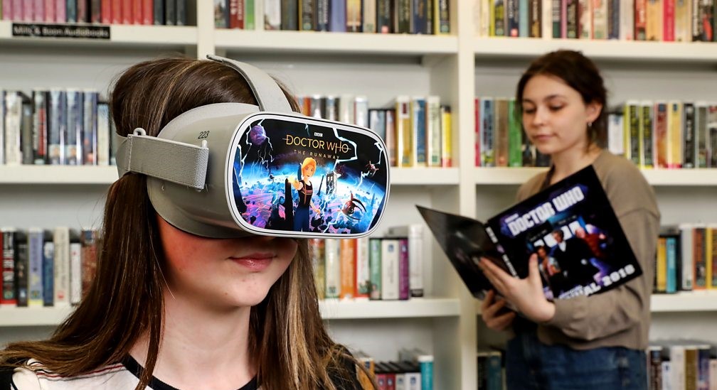 Girl watches Doctor Who on a VR headset in a library while another reads a book behind her