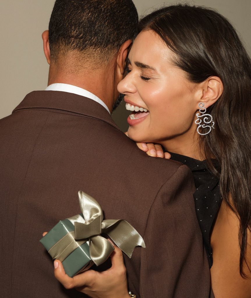A woman holds a small gift wrapped package and laughs, leaning on the shoulder of a brown-suited man