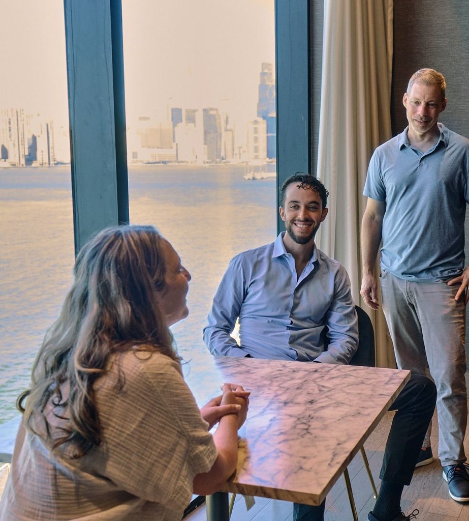 A woman sitting on a table by the window talking to two men