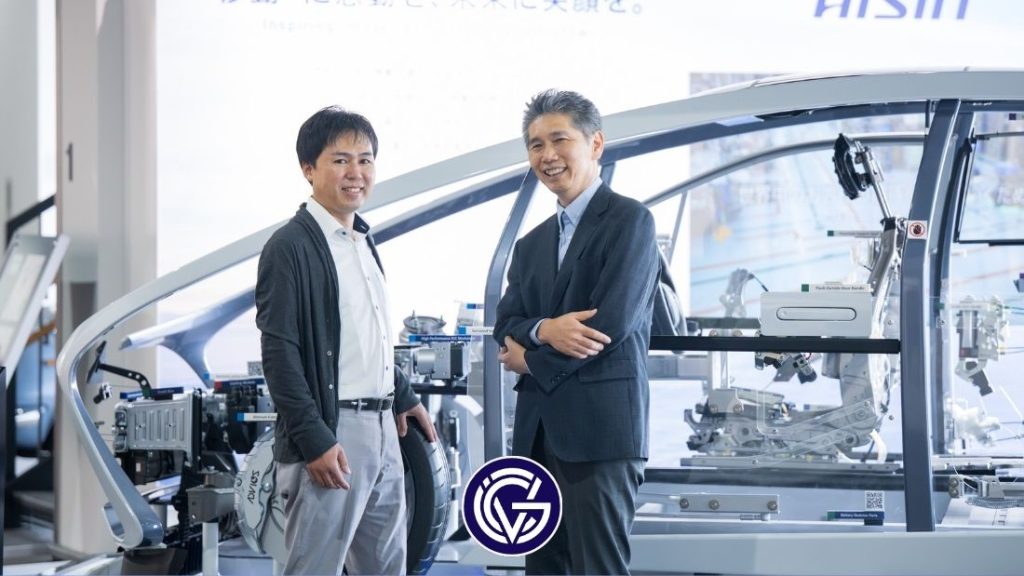 Two men stand in front of a car in a factory