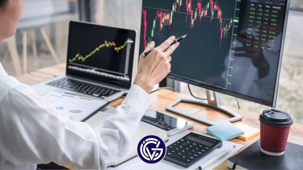 A woman sitting at a desk pointing a pen at one of two screens showing a share price pattern